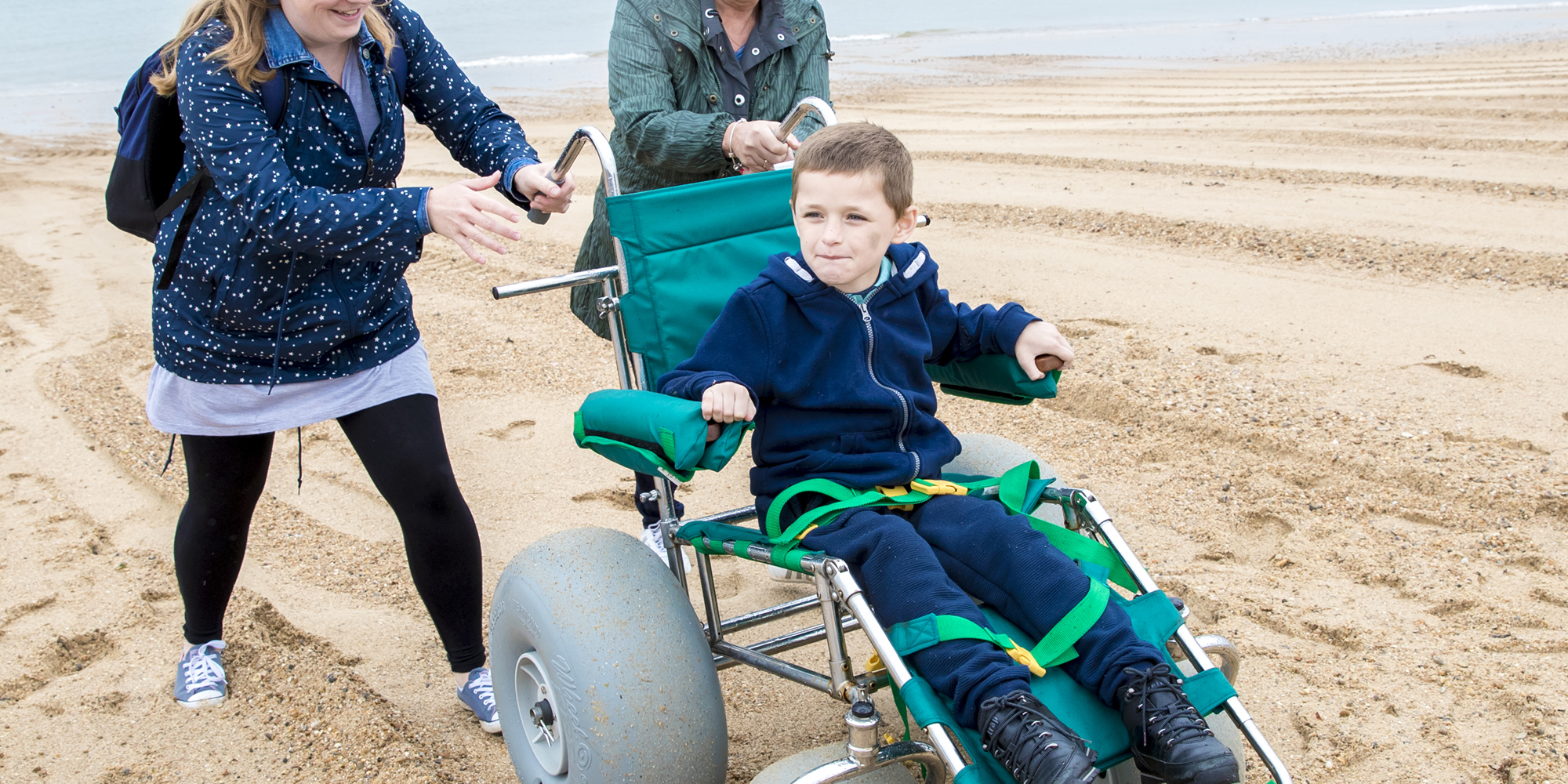 Two smiling women pushing a young boy in a wheelchair on a beach