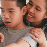 Young disabled boy looking at laptop with mum leaning over his shoulder pointing and smiling
