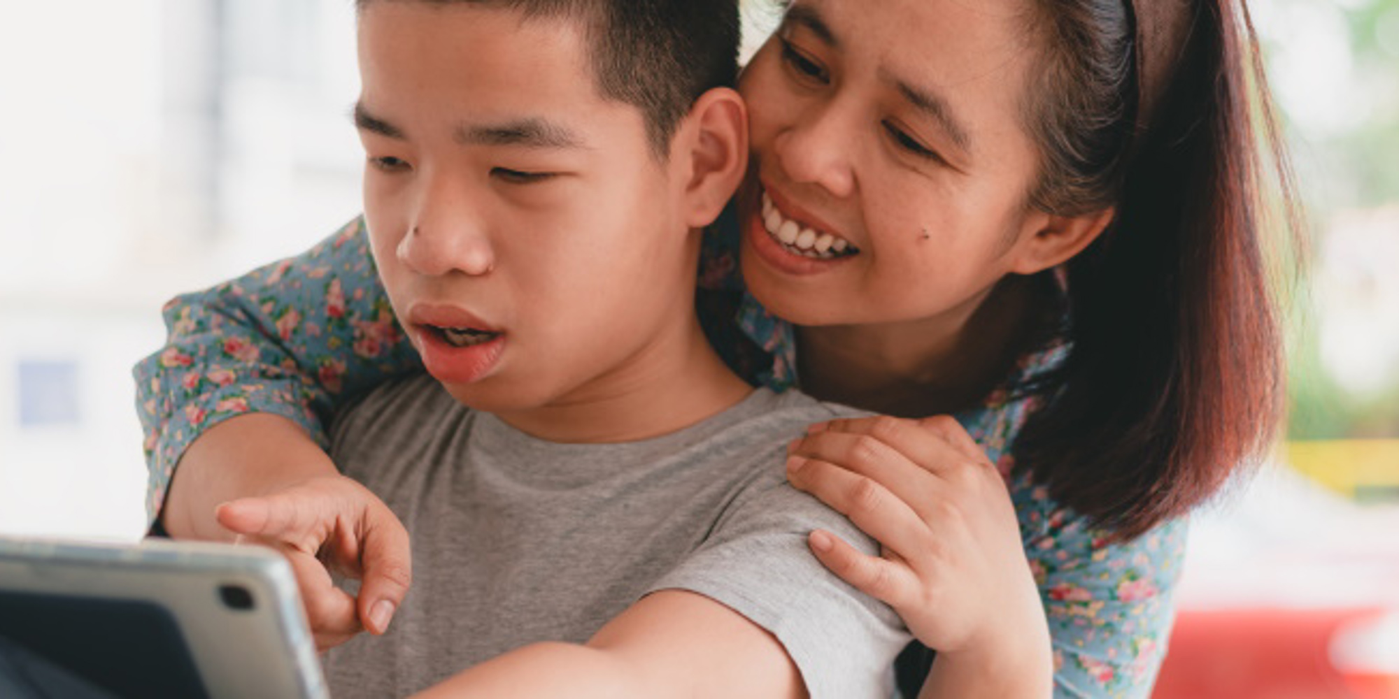 Young disabled boy looking at laptop with mum leaning over his shoulder pointing and smiling